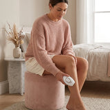 A woman sits on a pink velvet ottoman in a softly lit bedroom, using a white handheld device on her leg. She wears a chunky dusty pink sweater and cream shorts, with a bed, side table, and dried flowers in the background.