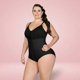 A woman modeling a black one-piece shapewear bodysuit with a front zipper and sheer mesh panel, set against a solid light pink studio background.
