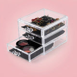 Clear acrylic makeup organizer with three drawers, shown open on a soft pink background. The organizer contains black makeup bags, two round blush compacts, and jewelry stored in the top compartment.