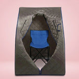 A portable quilted infrared sauna tent in dark gray with blue trim, shown with a blue folding chair inside on a solid pink background.